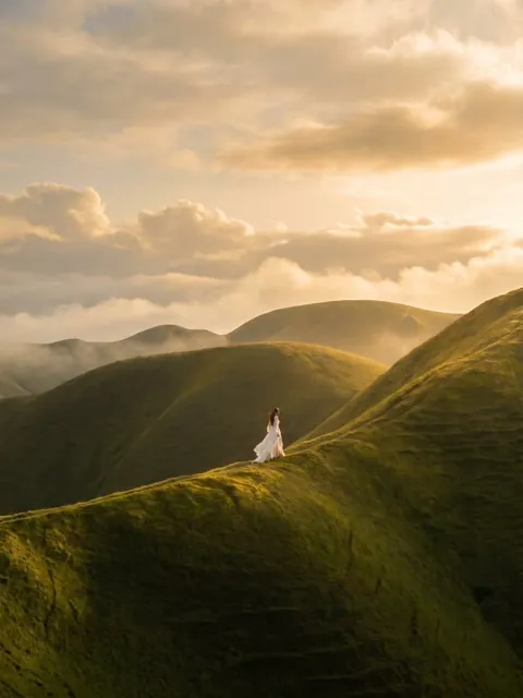Mujer de vestido blanco caminando sobre colinas verdes al atardecer